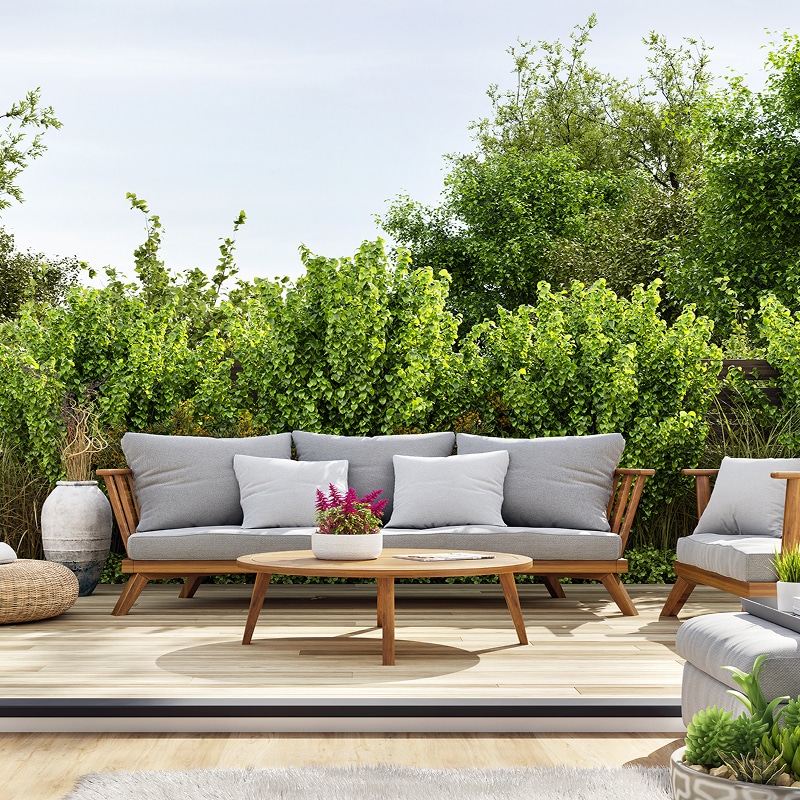 Salon de jardin moderne avec canapé et fauteuil en bois et coussins gris, sur une terrasse boisée, entouré d'une haie végétale dense.